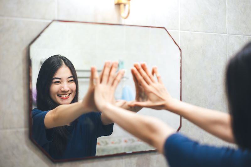 Image of a woman smiling at her reflection in the mirror, hands outstretched and touching the glass.