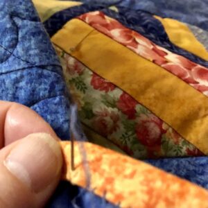 Fingers, needle, and thread on the edge of a colorful quilt shows hand-stitching the binding, the final step of making a quilt.