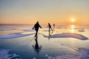 Image is of ice skaters on a frozen lake, skating toward he sunrise.